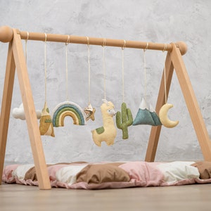 May include: A wooden baby play gym with a soft, padded mat and hanging toys. The toys include a rainbow, a star, a llama, a cactus, a mountain, and a crescent moon. The toys are made of felt and are in various shades of green, yellow, and blue.