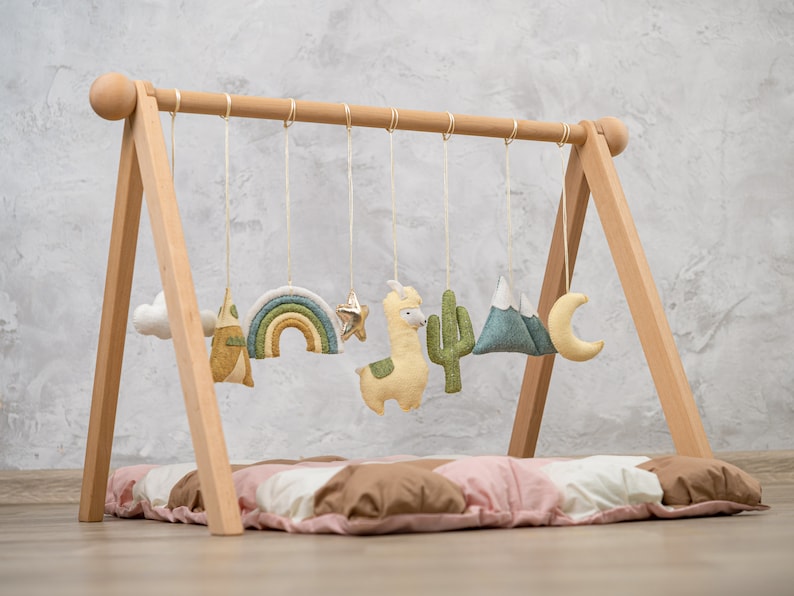 May include: A wooden baby play gym with a soft, padded mat and hanging toys. The toys include a rainbow, a star, a llama, a cactus, a mountain, and a crescent moon. The toys are made of felt and are in various shades of green, yellow, and blue.