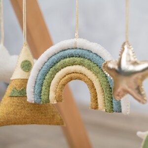 May include: A felt rainbow mobile with a white, green, and yellow color scheme. The rainbow is hanging from a white string. There is also a felt mountain and a gold star hanging from the mobile.