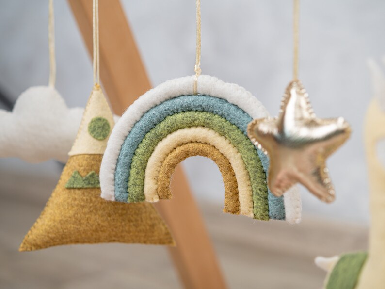 May include: A felt rainbow mobile with a white, green, and yellow color scheme. The rainbow is hanging from a white string. There is also a felt mountain and a gold star hanging from the mobile.