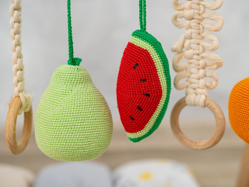 May include: A crocheted baby toy set with a green pear, a red watermelon, a wooden ring, and a white macrame ring.