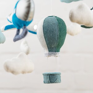 May include: A baby mobile with a blue airplane, white clouds, and a green and white hot air balloon with a basket. The mobile is made of felt and is hanging from a white ceiling.