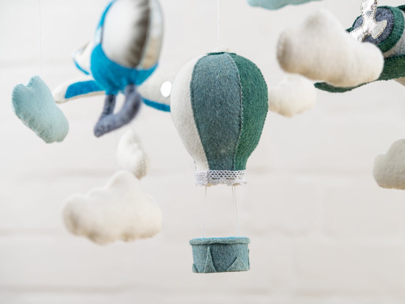 May include: A baby mobile with a blue airplane, white clouds, and a green and white hot air balloon with a basket. The mobile is made of felt and is hanging from a white ceiling.