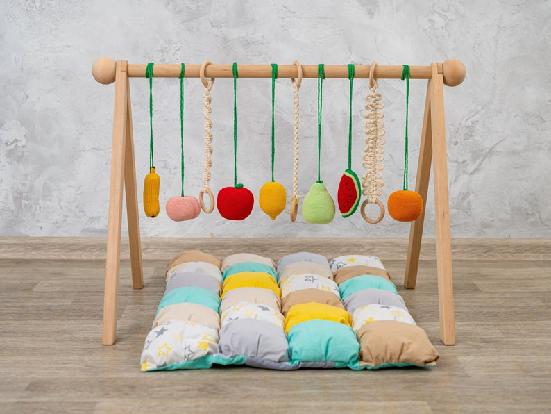 May include: A wooden baby play gym with a variety of hanging toys, including a banana, apple, pear, lemon, watermelon, and a knotted rope. The gym is set up on a colorful patchwork play mat.