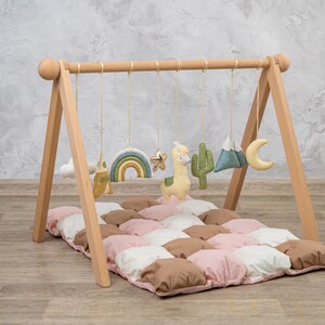May include: Wooden baby play gym with hanging toys including a rainbow, star, llama, cactus, mountains, and a crescent moon. The gym is set up on a pink, white, and brown quilted play mat.