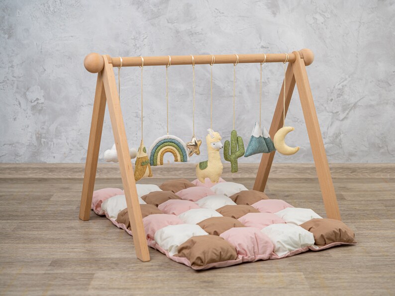 May include: Wooden baby play gym with hanging toys including a rainbow, star, llama, cactus, mountains, and a crescent moon. The gym is set up on a pink, white, and brown quilted play mat.