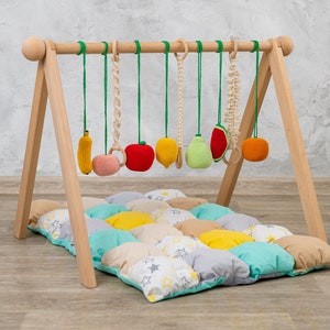 May include: Wooden baby play gym with a colorful fruit-themed hanging toy set. The gym has a padded play mat with a patchwork design of yellow, green, and gray squares with white stars.
