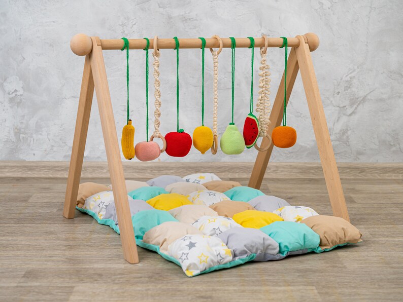 May include: Wooden baby play gym with a colorful fruit-themed hanging toy set. The gym has a padded play mat with a patchwork design of yellow, green, and gray squares with white stars.