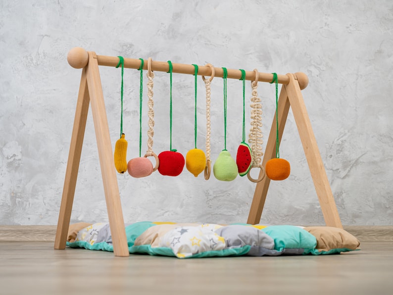 May include: A wooden play gym with a colorful assortment of hanging fruit-shaped toys, including a banana, apple, lemon, pear, watermelon, and orange. The gym is set up on a multi-colored padded play mat.