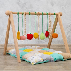 May include: A wooden play gym with a colorful fabric play mat and hanging fruit-shaped toys. The toys include a banana, apple, lemon, pear, watermelon, and orange.