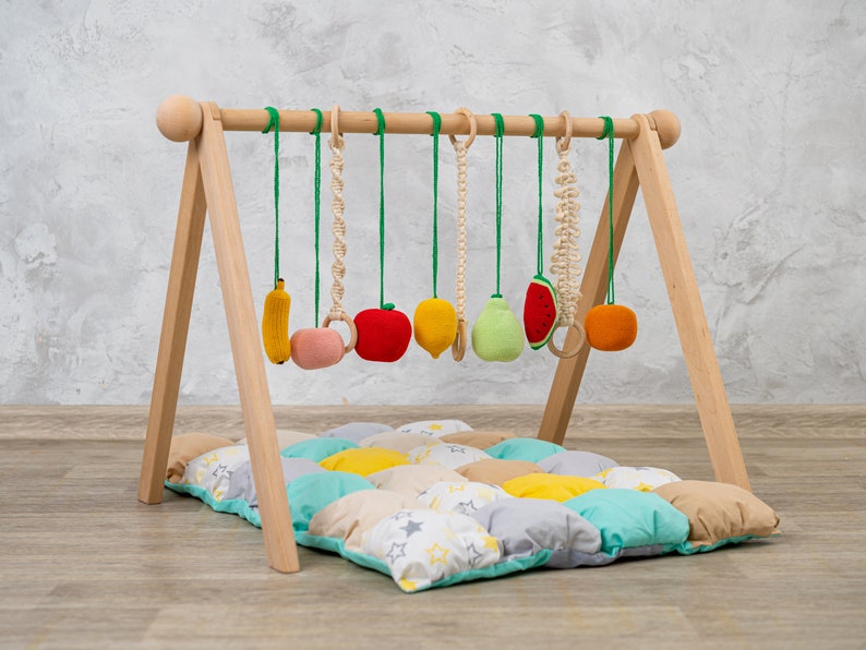 May include: A wooden play gym with a colorful fabric play mat and hanging fruit-shaped toys. The toys include a banana, apple, lemon, pear, watermelon, and orange.