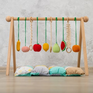 May include: Wooden play gym with hanging felt fruit toys, including a banana, apple, pear, watermelon, and orange. The gym is set up on a wooden floor with a colorful patterned cushion underneath.