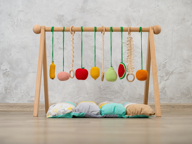 May include: Wooden play gym with hanging felt fruit toys, including a banana, apple, pear, watermelon, and orange. The gym is set up on a wooden floor with a colorful patterned cushion underneath.