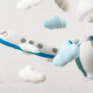 May include: A white and blue airplane, a green and white hot air balloon, and white and blue clouds hang from a mobile. The mobile is made of felt and is perfect for a baby's nursery.