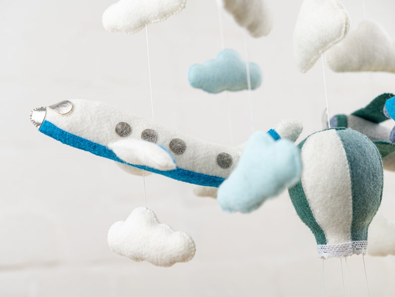 May include: A white and blue airplane, a green and white hot air balloon, and white and blue clouds hang from a mobile. The mobile is made of felt and is perfect for a baby's nursery.