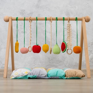 May include: A wooden baby play gym with a variety of colorful, crocheted fruit and vegetable toys hanging from it. The gym is set up on a light wood floor with a multi-colored pillow underneath.
