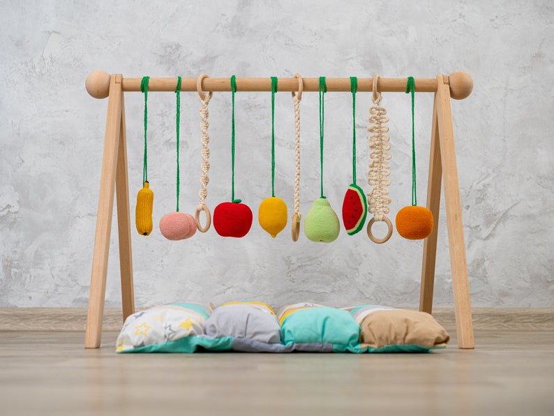 May include: A wooden baby play gym with a variety of colorful, crocheted fruit and vegetable toys hanging from it. The gym is set up on a light wood floor with a multi-colored pillow underneath.