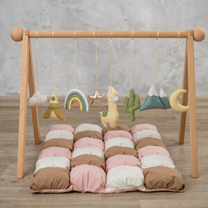 May include: A wooden play gym with a hanging mobile featuring a rainbow, a star, a llama, a cactus, a mountain range, a cloud, and a crescent moon. The gym is set up on a brown, pink, and white quilted play mat.
