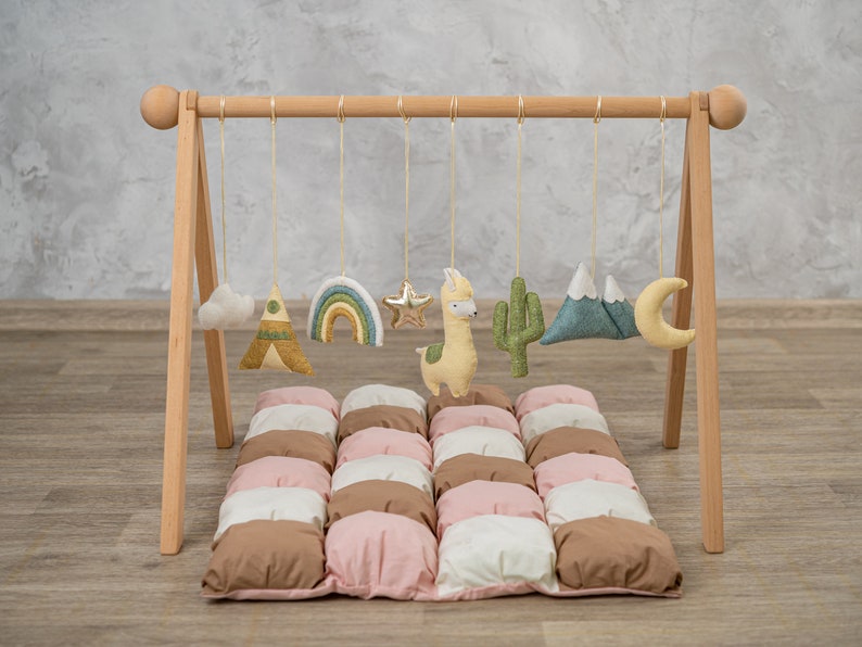 May include: A wooden play gym with a hanging mobile featuring a rainbow, a star, a llama, a cactus, a mountain range, a cloud, and a crescent moon. The gym is set up on a brown, pink, and white quilted play mat.