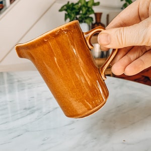 Puede incluir: Un creamer de cerámica marrón con asa. El creamer está sobre una superficie de mármol blanco.