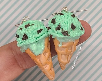 May include: A pair of mint green ice cream cone earrings with chocolate chips, hanging from silver earring hooks. The ice cream is melting down the cone.