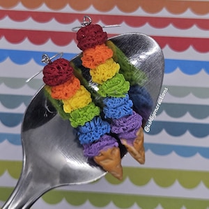 May include: A pair of handmade earrings in the shape of ice cream cones. Each earring has a rainbow of colours: red, orange, yellow, green, blue, and purple. The earrings are displayed on a silver spoon.