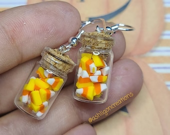 May include: A pair of earrings featuring miniature glass jars filled with yellow, orange, and white candy corn. The jars have cork lids and are attached to silver earring hooks. The earrings are held in a hand against a striped background.