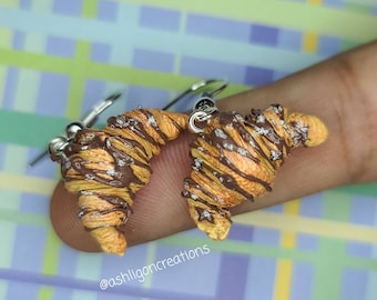 Pode incluir: Um par de brincos de croissant com cobertura de chocolate e açúcar em pó. Os brincos são feitos de argila polimérica e têm ganchos prateados.