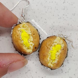 Op de afbeelding: Een paar oorbellen in de vorm van gebakken aardappelen met gele boter en kaas. De oorbellen zijn gemaakt van polymer clay en zijn bevestigd aan zilveren haken.
