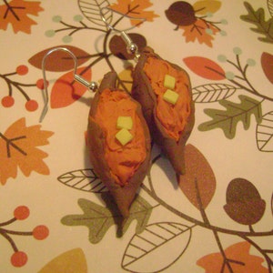 May include: A pair of earrings shaped like sweet potatoes with a light orange glaze and a small square of white fondant on top. The earrings are hanging from silver hooks.