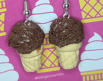 May include: A pair of dangle earrings featuring a chocolate ice cream cone with sprinkles on top. The earrings are made of polymer clay and have silver hooks. The background is a pink and yellow pattern of ice cream cones.