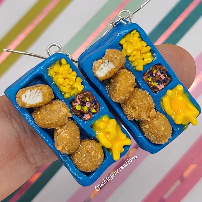 May include: A pair of blue tray earrings with a miniature depiction of a chicken nugget meal. The tray has four compartments filled with chicken nuggets, mac and cheese, corn, and a brown sauce. The earrings are made of polymer clay and have silver hooks.
