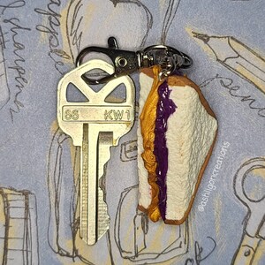 May include: A key chain featuring a miniature peanut butter and jelly sandwich with purple jelly and a gold peanut butter filling. The sandwich is attached to a silver key with the numbers "66" and "KW1" on it.