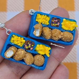 May include: A pair of blue tray earrings with a miniature school lunch meal. The tray has four compartments with chicken nuggets, corn, tater tots, and a side of gravy. The earrings are made of polymer clay and have silver hooks.