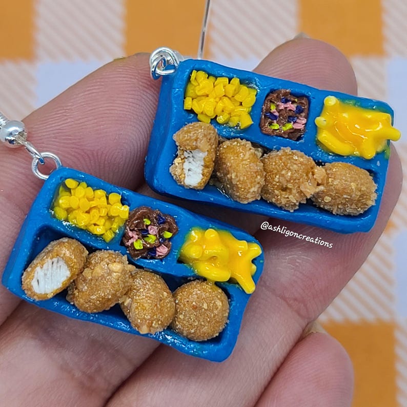 May include: A pair of blue tray earrings with a miniature school lunch meal. The tray has four compartments with chicken nuggets, corn, tater tots, and a side of gravy. The earrings are made of polymer clay and have silver hooks.