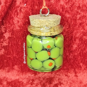May include: A clear glass jar filled with green olives with orange pimentos. The jar has a cork stopper and a metal ring for hanging. The background is a textured red fabric. The text "@ashligoncreations" is on the side of the jar.