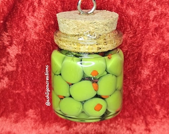 Può includere: Un piccolo barattolo di vetro pieno di olive verdi con peperoni arancioni. Il barattolo ha un tappo di sughero e un anello di metallo per appenderlo. Lo sfondo è un tessuto rosso strutturato. Il testo "@ashligoncreations" è sul lato del barattolo.