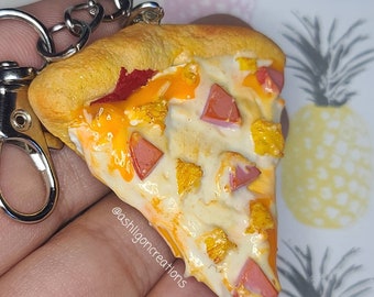 May include: A miniature pizza slice keychain with a yellow crust, red sauce, white cheese, and yellow and orange toppings. The keychain is held in a hand and has a silver chain attached. The text '@ashligoncreations' is visible on the keychain.