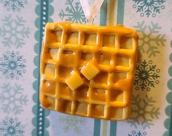 May include: A yellow waffle ornament with a brown glaze and white string for hanging. The waffle has a square shape and is decorated with small squares.