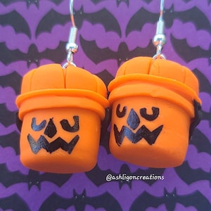 Puede incluir: Un par de pendientes de color naranja en forma de calabaza con caras de calabaza pintadas de negro. Los pendientes están sobre un fondo morado con siluetas de murciélagos. Los pendientes están hechos a mano y tienen ganchos plateados. @ashligoncreations