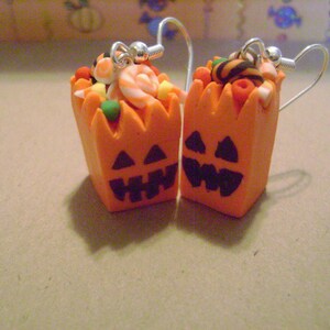 May include: A pair of orange pumpkin-shaped earrings with candy inside. The earrings are made of polymer clay and have a black jack-o'-lantern face.
