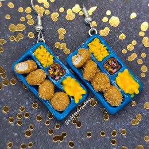 May include: A pair of blue tray-shaped earrings with a miniature depiction of a school lunch tray filled with chicken nuggets, mac and cheese, and corn. The earrings are made of polymer clay and have silver hooks.