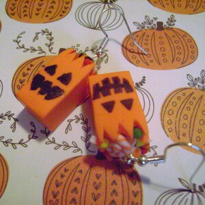 May include: A pair of orange pumpkin-shaped earrings with black painted faces. The earrings are made of polymer clay and have silver hooks.