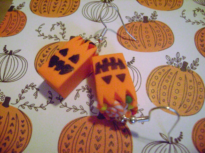 May include: A pair of orange pumpkin-shaped earrings with black painted faces. The earrings are made of polymer clay and have silver hooks.