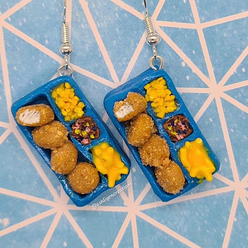 May include: A pair of blue and white earrings shaped like a miniature plastic tray of chicken nuggets, corn, and mac and cheese. The earrings are made of polymer clay and have silver hooks.