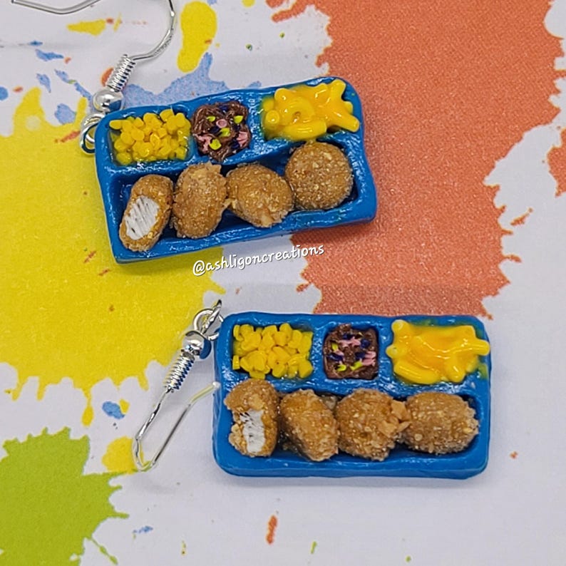 May include: A pair of blue and white school lunch tray earrings with a silver hook. The tray has four chicken nuggets, corn, a side of mystery meat, and tater tots.