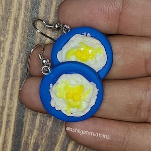Puede incluir: Un par de pendientes de arcilla polimérica azul y blanca con forma de platos de comida. La comida es blanca con detalles amarillos y parece arroz con salsa amarilla y un cuadrado amarillo en la parte superior. Los pendientes están en un gancho plateado.