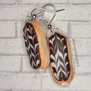 May include: A pair of dangle earrings featuring a chocolate eclair with white icing. The eclairs are made of polymer clay and have a realistic appearance.
