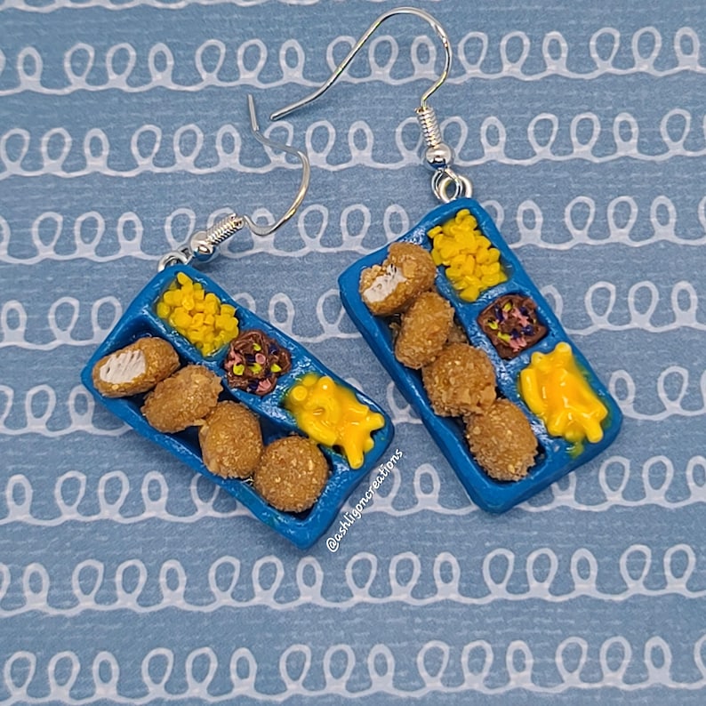 May include: A pair of blue tray earrings with a miniature depiction of a school lunch tray filled with chicken nuggets, mac and cheese, and fruit cocktail. The earrings are made of polymer clay and have silver hooks.