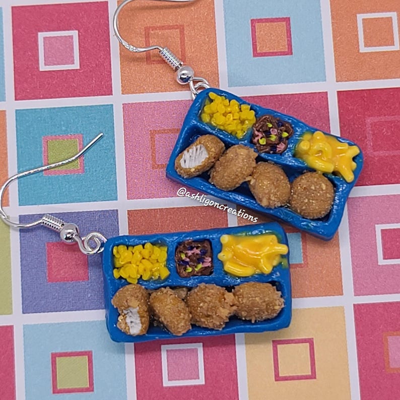 May include: A pair of blue and yellow school lunch tray earrings with a white chicken nugget, corn, and fries. The earrings are made of polymer clay and have silver hooks. The earrings are handmade and feature the text "@ashligoncreations".